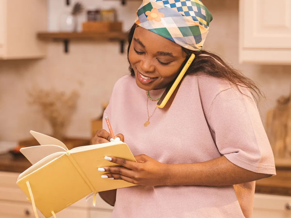 A woman in her kitchen, on the phone, jotting in a notebook